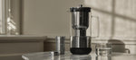 Aarke Purifier placed on a marble table with its stainless steel filter container and a glass of water beside it.