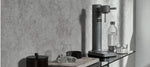 Aarke Carbonator 3 in Hammertone placed against a textured grey wall, next to a glass and a PET bottle of sparkling water.