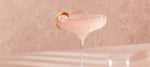 Close-up of a coupe glass with a pale pink cocktail, a large ice cube, and a grapefruit twist garnish.