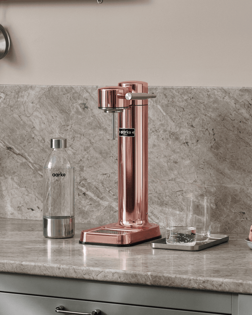 Aarke Carbonator 3 in Copper displayed on a marble kitchen countertop, alongside a PET water bottle and two drinking glasses.