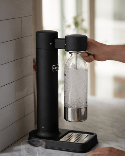 Aarke Carbonator 3 in Matte Black carbonating sparkling water in a PET bottle on a marble countertop, with a hand operating the lever.