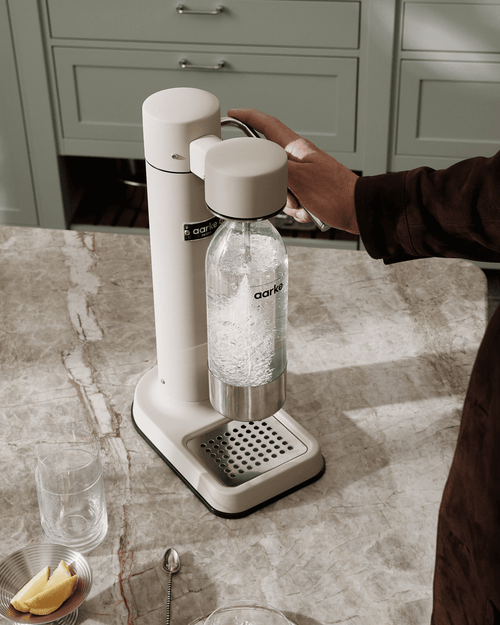 Aarke Carbonator 3 in Sand carbonating sparkling water in a PET bottle on a marble countertop, alongside a drinking glass and lemon wedges.