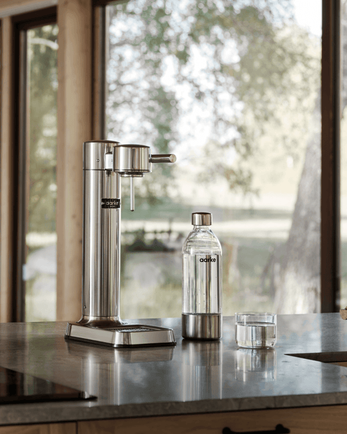 Aarke Carbonator 3 in Stainless Steel displayed on a kitchen countertop alongside a water bottle and a glass of sparkling water.