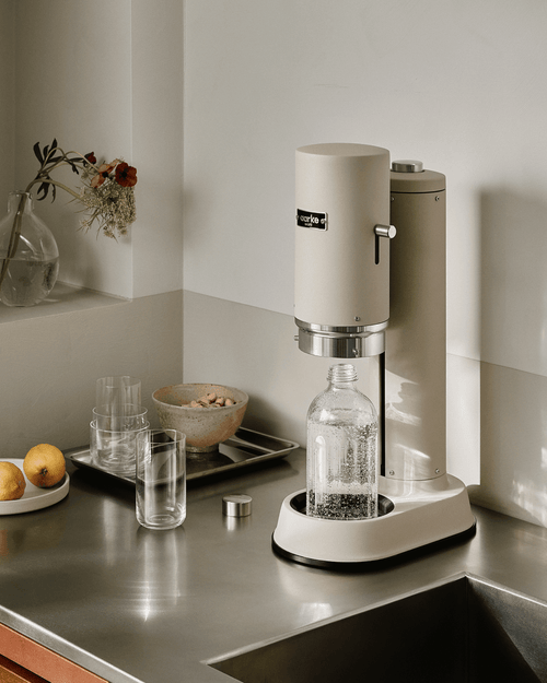 Aarke Carbonator Pro sparkling water maker in Sand with glass bottle on a stainless steel kitchen countertop.