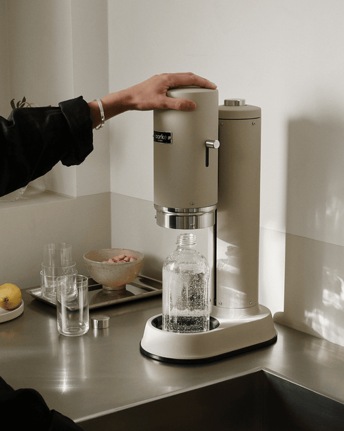 Aarke Carbonator Pro in Sand with a glass water bottle, showing the push-down carbonation mechanism being operated by hand.