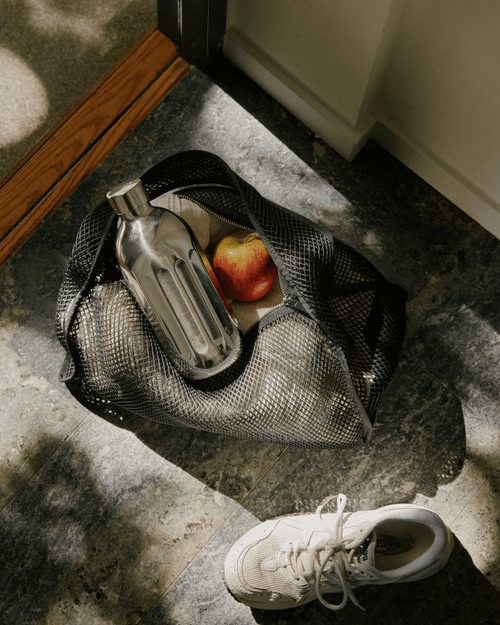 Aarke To-Go Bottle Large in stainless steel, placed inside a black mesh gym bag alongside an apple, with a white sneaker nearby.