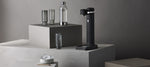 Aarke Carbonator 3 Matte Black displayed on a minimalist grey pedestal alongside a PET water bottle, ribbed drinking glasses, and a bowl of ice cubes.