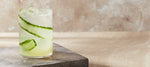 A refreshing cocktail with ice and cucumber served in a glass on a marble slab against a soft beige background.