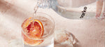 A close-up of sparkling water being poured into a glass filled with ice and a slice of blood orange, on a pink marble surface.