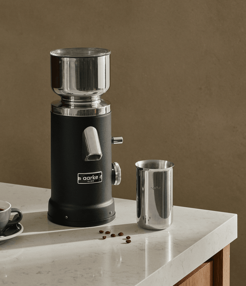 Aarke Coffee Grinder in Matte Black with stainless steel grounds container and coffee beans on a kitchen countertop.