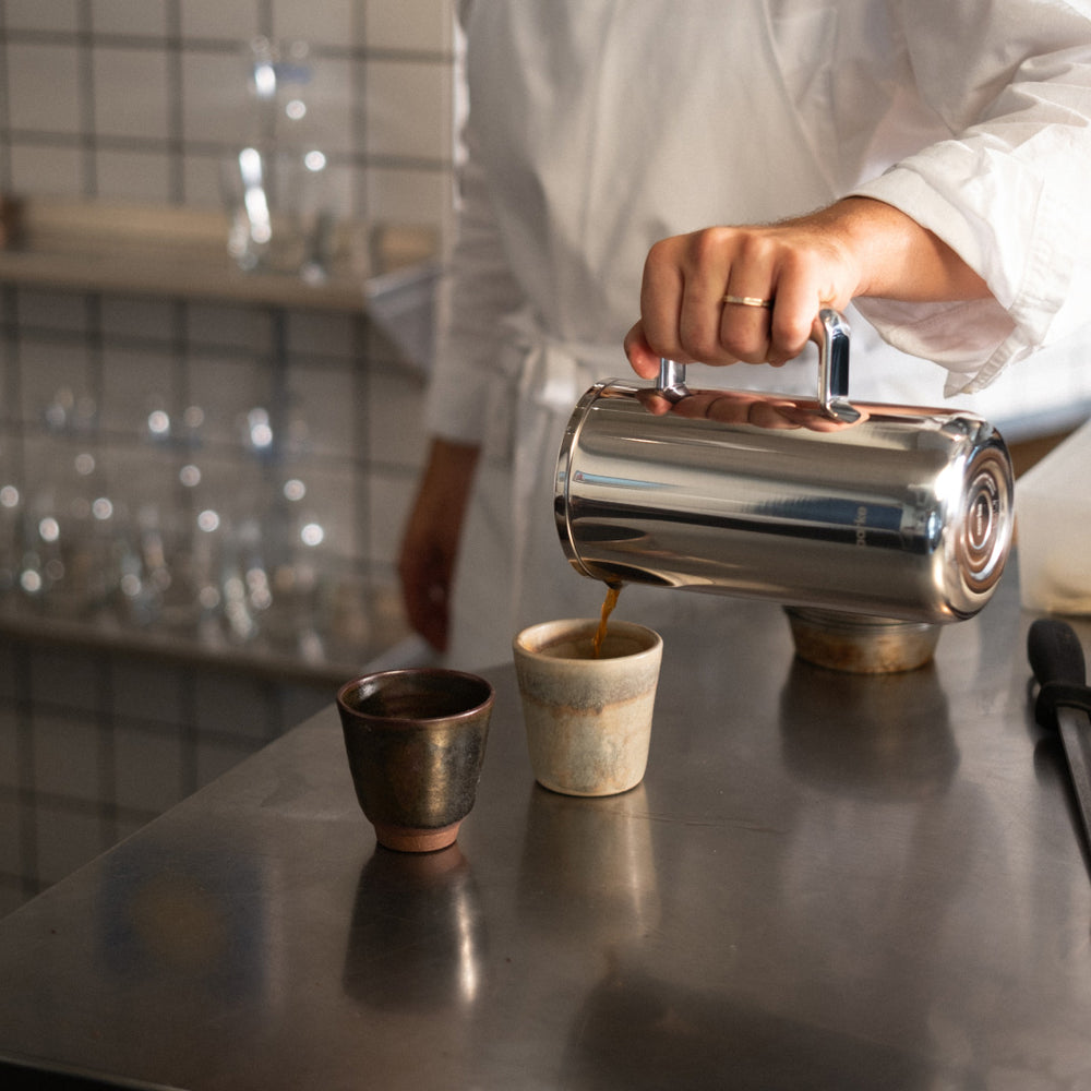 Person pouring coffee from a steel Coffee Press into a ceramic cup on a kitchen counter.