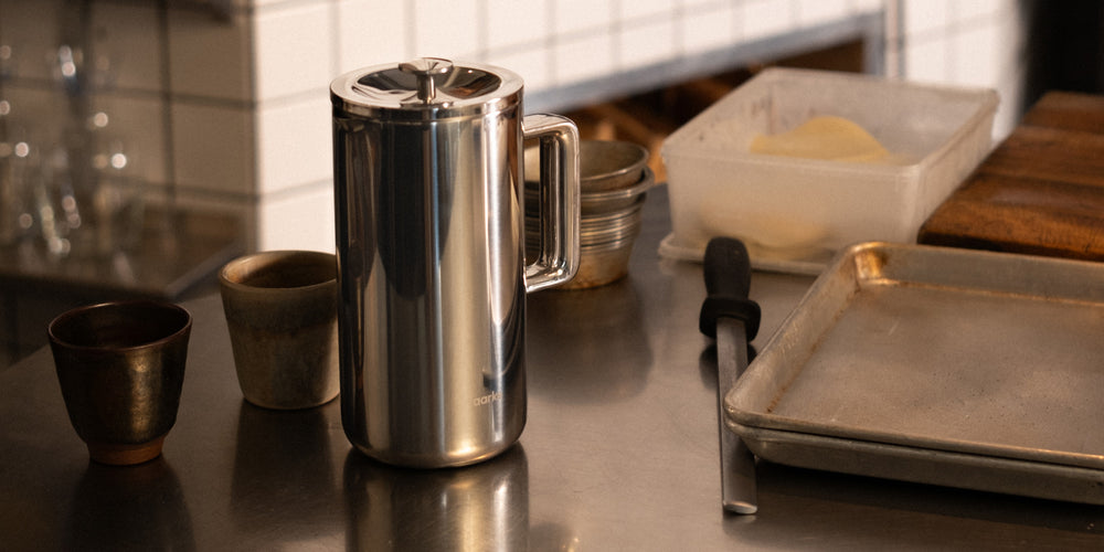 Stainless steel coffee press on a kitchen counter with various items including a tray and bowls.