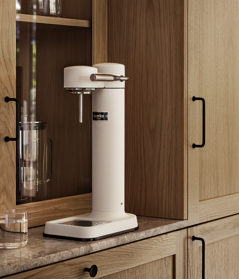 Aarke Carbonator 3 in matte white finish on a kitchen counter with wooden cabinets, next to a glass of sparkling water.