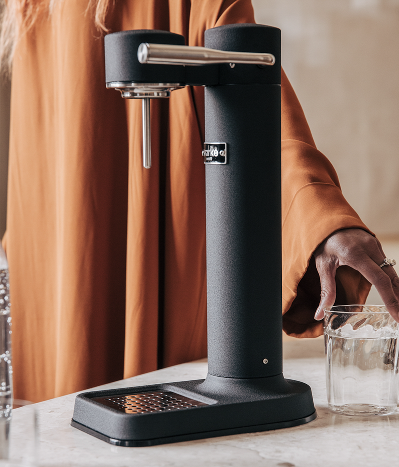 Close-up of the Aarke Carbonator 3 in matte black, with a person placing a glass of sparkling water beside it.