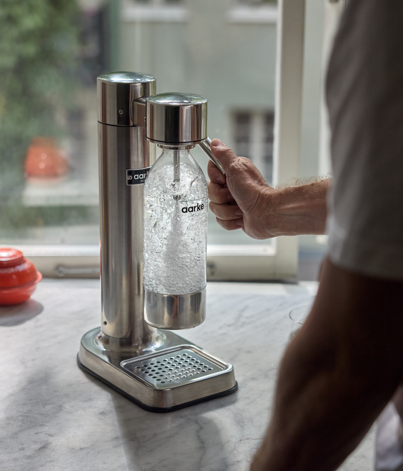 Person using the Aarke Carbonator 3 in stainless steel, carbonating water in a reusable pet water bottle on a marble countertop.