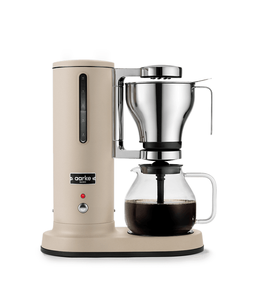 Aarke sand-colored automatic pour-over coffee maker with stainless steel filter brewing coffee into glass carafe.