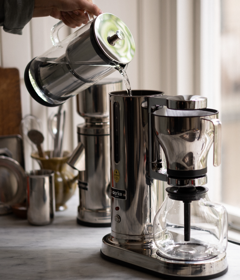 Person pouring water into the Aarke stainless steel coffee maker, with the Aarke coffee grinder in the background.