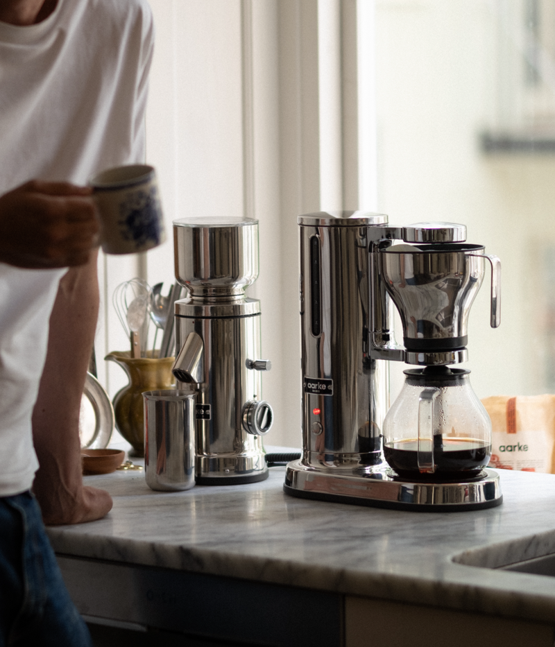 Person holding a coffee mug beside the Aarke stainless steel coffee maker and grinder, with freshly brewed coffee in the glass carafe.