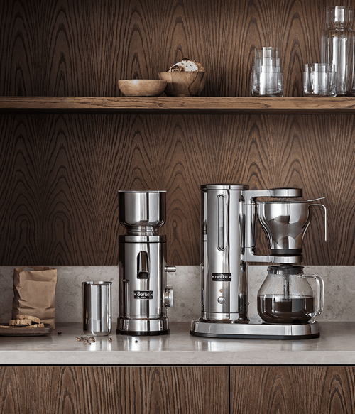 Aarke Coffee Station featuring the Coffee Grinder and Coffee Maker side by side on a kitchen counter with warm wooden cabinets and shelves in the background.