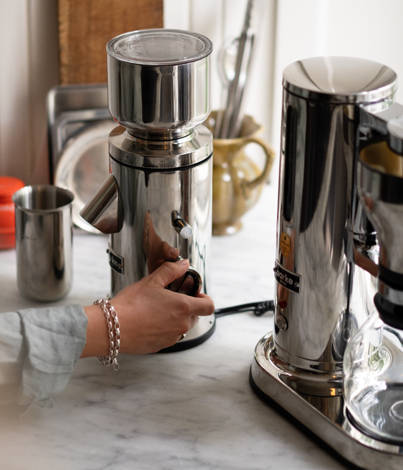 Close-up of a hand adjusting the grind setting on the Aarke stainless steel coffee grinder, with the Aarke stainless steel coffee maker beside it.