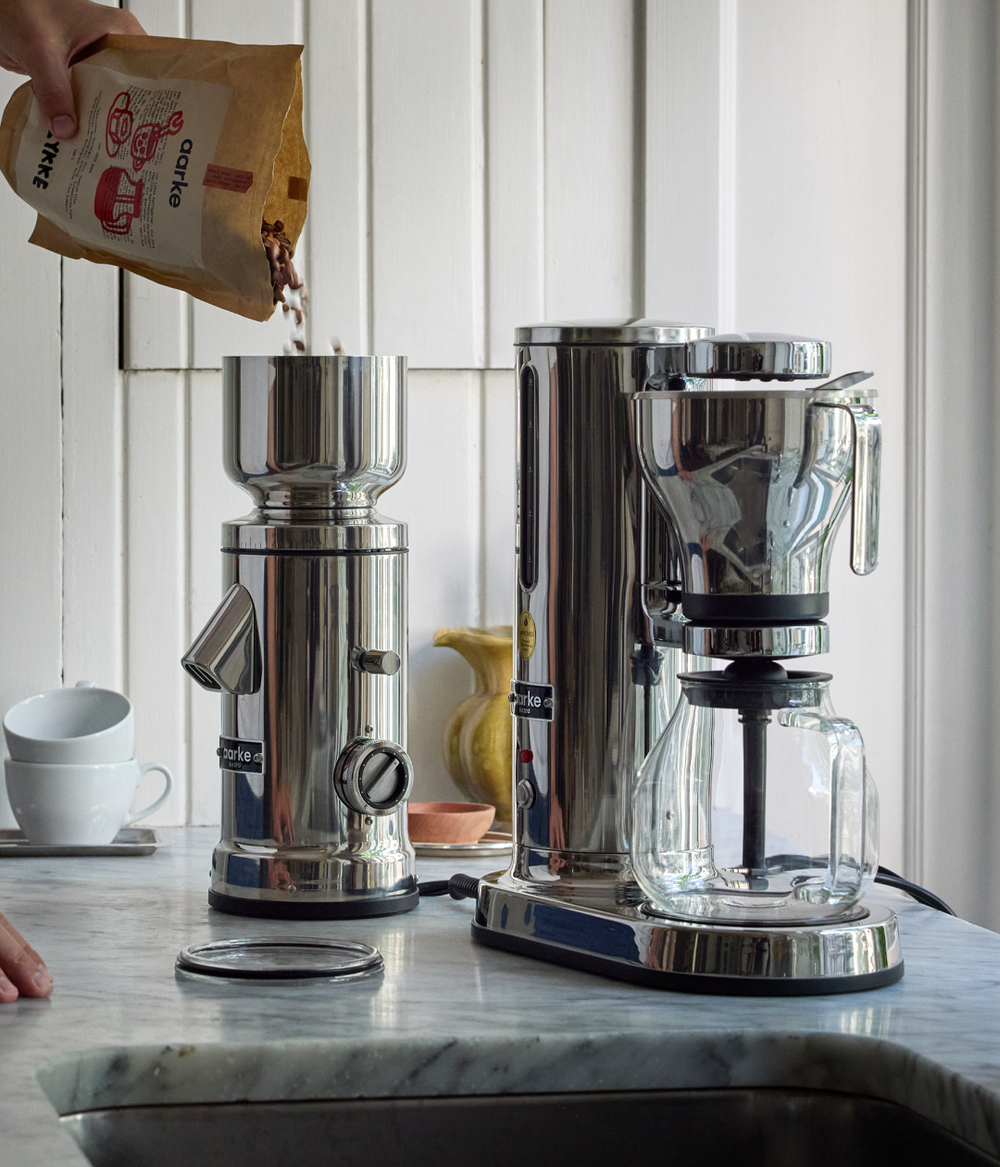 Aarke stainless steel coffee grinder with coffee beans being poured in, next to the Aarke stainless steel coffee maker on a marble countertop.