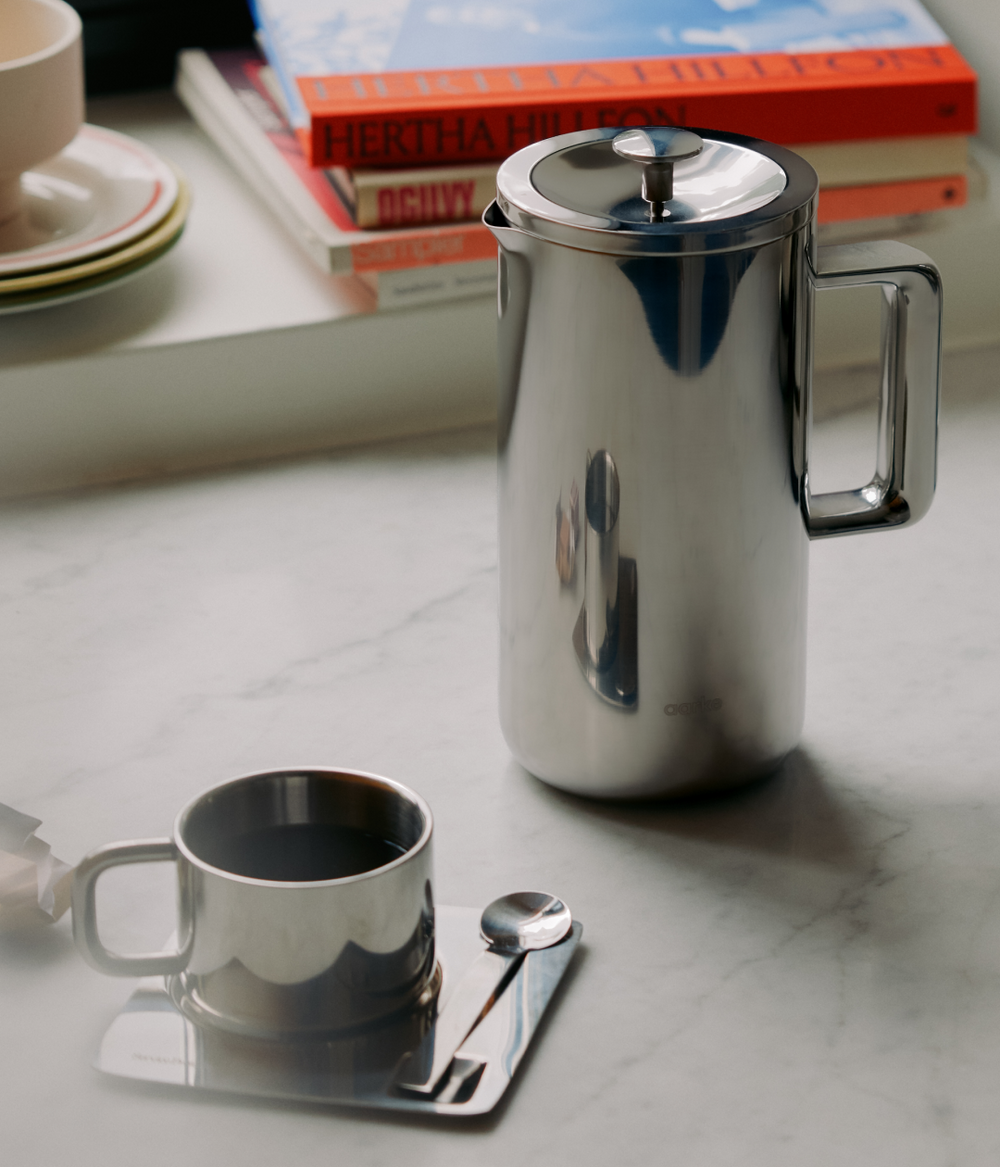 Aarke stainless steel French Press with matching cup and spoon on marble table.