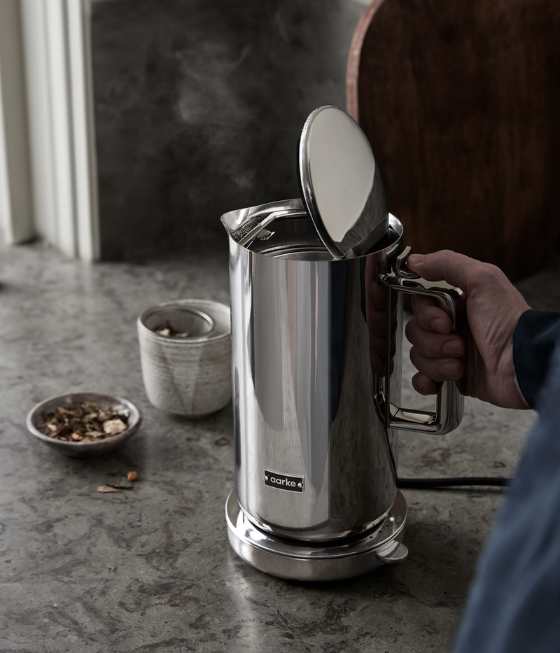A person opening the lid of a steaming Aarke kettle in stainless steel in a kitchen setting with tea ingredients.