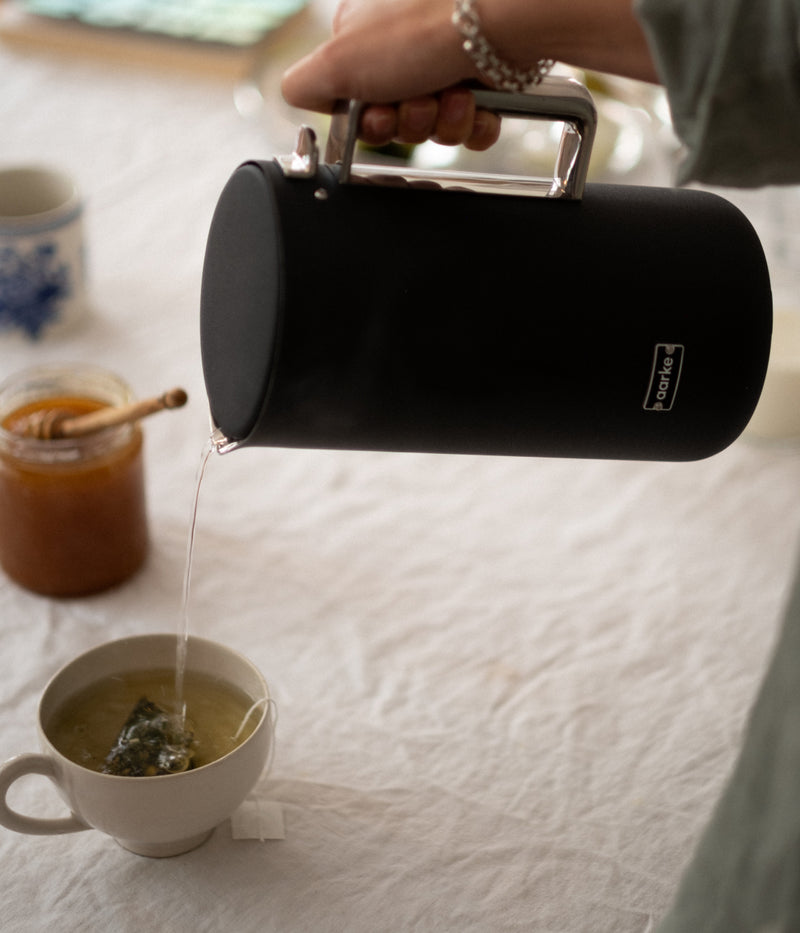 Person pouring hot water from the Aarke matte black electric kettle into a teacup with a tea bag