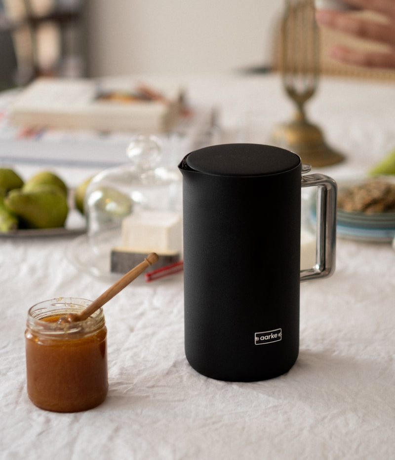 Aarke matte black electric kettle on a breakfast table next to a jar of honey and fresh fruit.