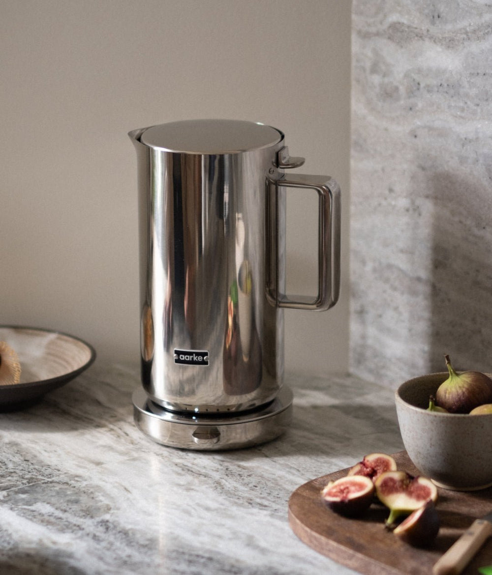 Stainless steel coffee press on a marble countertop with figs and a bowl.