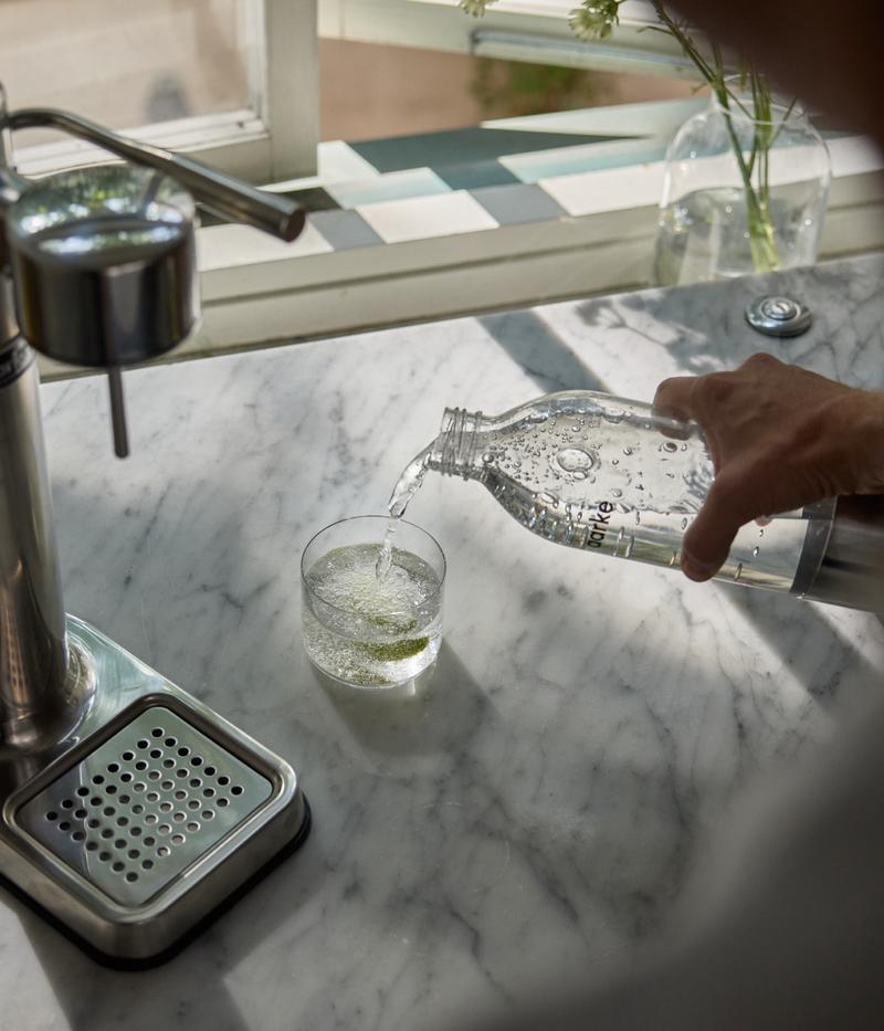 Person pouring sparkling water from an Aarke PET bottle into a glass, with the Aarke Carbonator 3 in stainless steel on the counter.