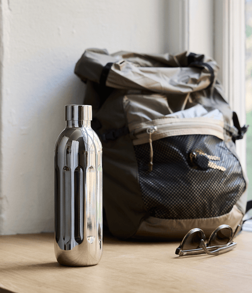 Aarke Stainless Steel Bottle Small placed on a wooden surface next to a backpack and sunglasses by a window.