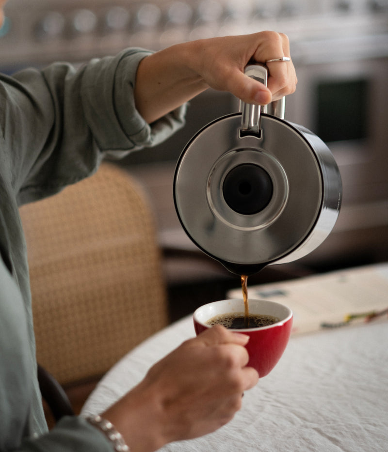 Close-up of a hand pouring coffee from the Aarke stainless steel thermal jug into a red coffee cup.
