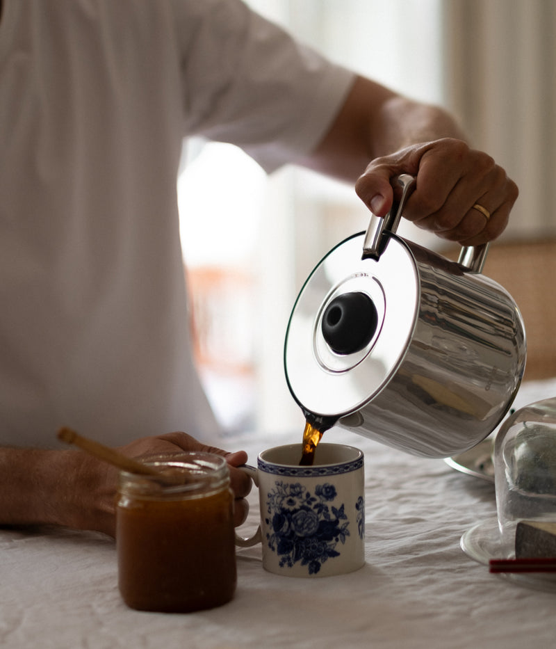 Person pouring coffee from the Aarke stainless steel thermal jug into a floral porcelain mug, with a jar of jam on the table.