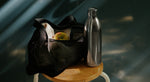 Black mesh bag with an apple, to-go water bottle in stainless steel on a wooden stool against a blurred background.