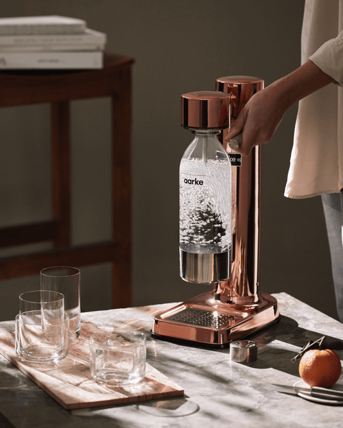 Aarke Carbonator 3 in Copper carbonating sparkling water in a PET bottle on a marble countertop, alongside drinking glasses and an orange.