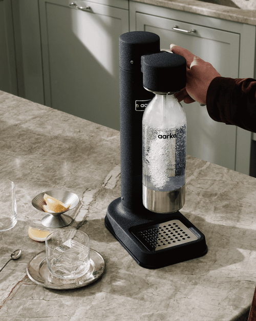 Aarke Carbonator 3 in Matte Black carbonating sparkling water in a PET bottle on a marble countertop, alongside a glass of water and lemon wedges.