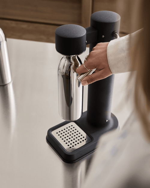 Aarke Carbonator 3 in Matte Black with a stainless steel water bottle, showing a hand operating the carbonation lever on a kitchen countertop.