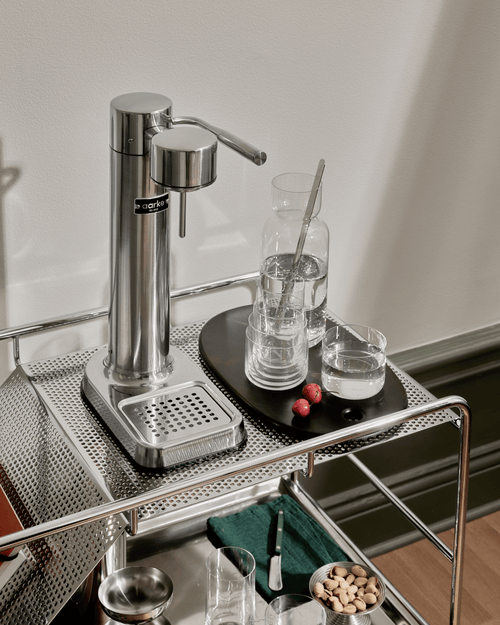 Aarke Carbonator 3 in Stainless Steel displayed on a stainless steel serving trolley, alongside nesting glasses, a water carafe and fresh cherries.