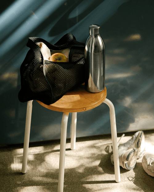 Aarke To-Go Bottle Large in stainless steel, standing on a wooden stool beside a black gym bag and white sneakers.