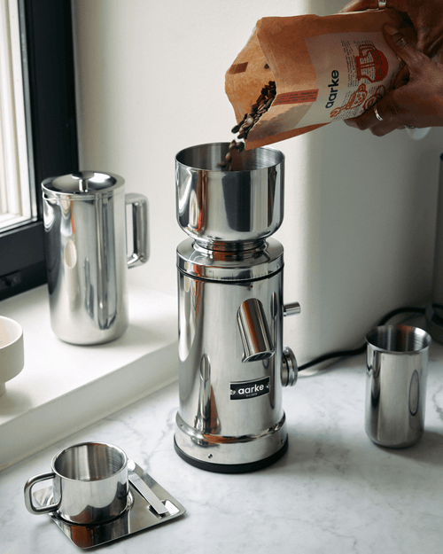 Whole coffee beans being poured into the stainless steel hopper of the Aarke Coffee Grinder, an electric flat burr coffee grinder with 60 grind settings.