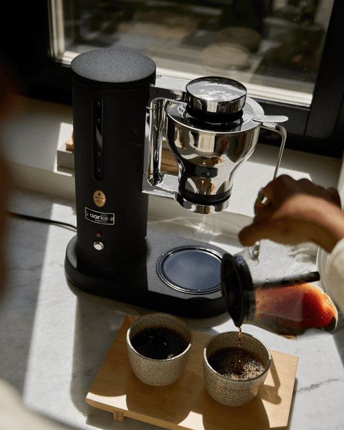 Freshly brewed filter coffee being poured into two ceramic cups from the glass carafe of the Aarke Coffee Maker in Matte Black.