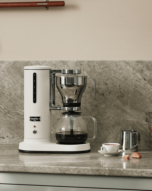 Aarke Coffee Maker in Matte White with glass carafe, stainless steel filter basket and freshly brewed coffee on a marble countertop.
