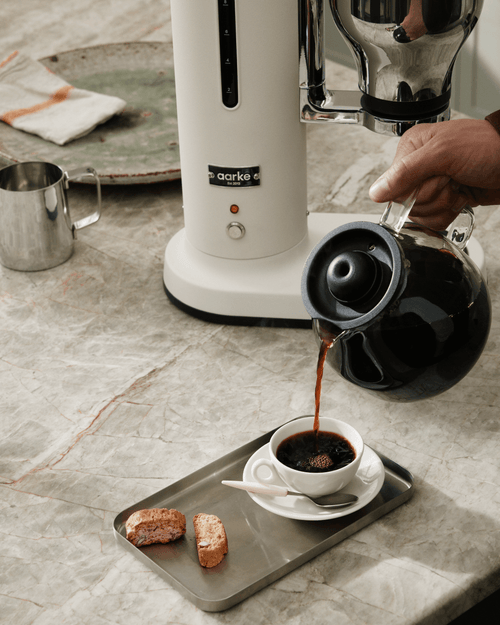 Freshly brewed coffee being poured from the glass carafe of the Aarke Coffee Maker in Matte White into a coffee cup.