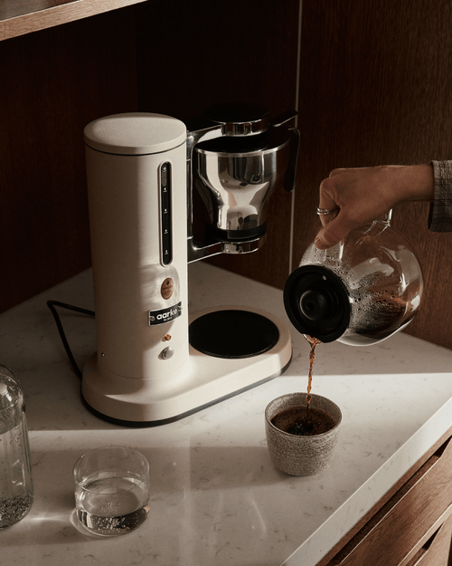Freshly brewed filter coffee being poured from the glass carafe of the Aarke Coffee Maker in Sand into a ceramic cup.