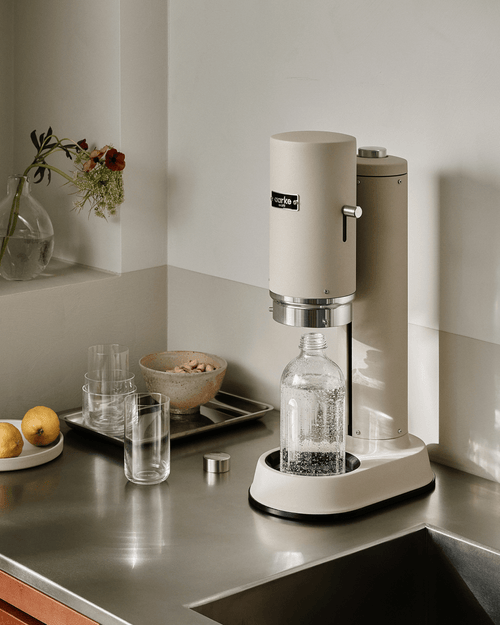 Aarke Carbonator Pro sparkling water maker in Sand with glass bottle on a stainless steel kitchen countertop.