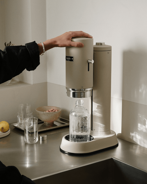 Aarke Carbonator Pro in Sand with a glass water bottle, showing the push-down carbonation mechanism being operated by hand.