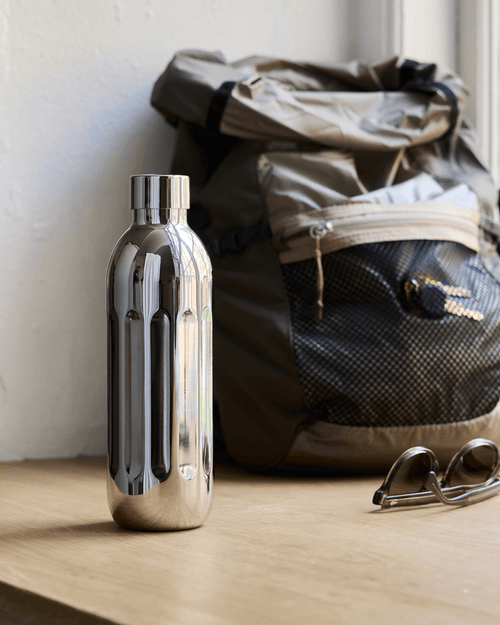 Aarke To-Go Bottle Small in stainless steel, standing on a wooden surface beside a backpack and sunglasses.