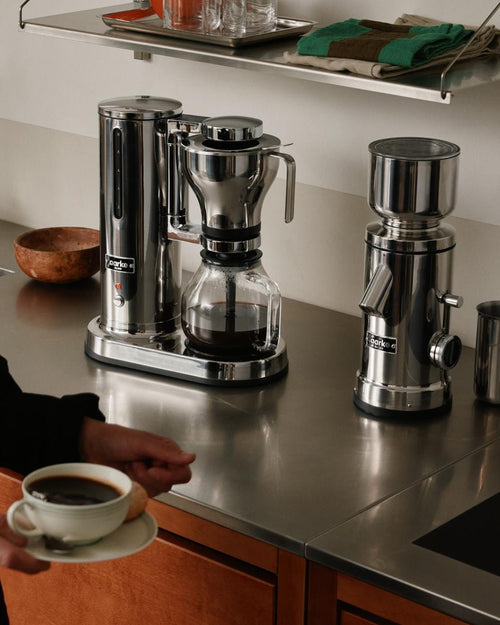 Aarke Coffee Maker and Coffee Grinder in stainless steel displayed side by side on a kitchen countertop, with freshly brewed coffee in a glass carafe.