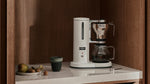Aarke Coffee Maker in Matte White brewing filter coffee in a warm wood-paneled kitchen with a marble countertop.